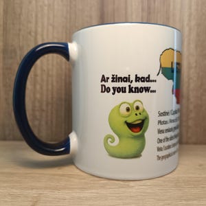 Puede incluir: Taza de cerámica blanca con asa y borde azules. La taza presenta un gusano de dibujos animados verde y texto en inglés y otro idioma. La taza está sobre una superficie de madera.