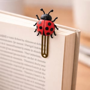 May include: A red and black ladybug bookmark is attached to the pages of a book. The ladybug has black spots and a black head. The bookmark is made of metal and is gold in color. The book has beige pages.