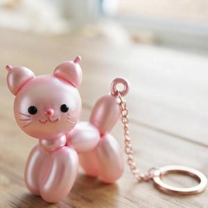 May include: A light pink cat-shaped keychain with a metallic finish, designed to resemble a balloon animal. The figure features small ears, black eyes, and a simple drawn face, attached to a matching pink metal chain and a circular key ring.