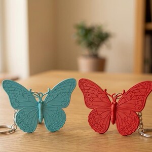 Op de afbeelding: Twee vlindervormige sleutelhangers, één in blauwgroen en één in rood, elk met ingewikkelde reliëfpatronen op de vleugels en een zilverkleurige metalen ring aan de zijkant, naast elkaar geplaatst op een plat oppervlak.
