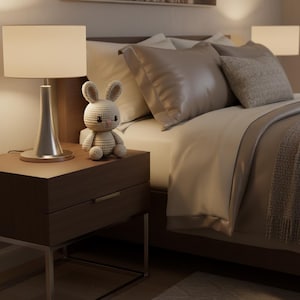 Puede incluir: Escena de dormitorio con una mesita de noche con una l&aacute;mpara plateada y un conejito de ganchillo. La cama tiene ropa de cama y almohadas de tonos neutros. La habitaci&oacute;n tiene un ambiente c&aacute;lido y acogedor, con iluminaci&oacute;n suave.