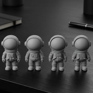 Pode incluir: Quatro estatuetas de astronautas cinzentos em fila. Cada estatueta tem um capacete arredondado, um fato espacial detalhado e botas. As estatuetas estão numa superfície escura, com uma caneta e um caderno no fundo.