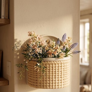 May include: A beige ceramic wall-mounted basket planter filled with pink, purple, and white flowers. The basket has a woven texture and a white rim. The planter is mounted on a beige wall.