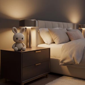 Puede incluir: Un ambiente de dormitorio con un conejito de ganchillo sobre una mesita de noche de madera oscura. Una l&aacute;mpara moderna con acabado met&aacute;lico est&aacute; sobre la mesa. La cama tiene almohadas y una manta. La habitaci&oacute;n est&aacute; suavemente iluminada.