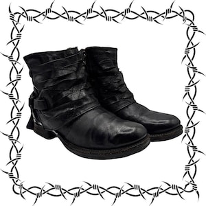 May include: A pair of black ankle boots featuring a textured, ruched upper design with a side strap and buckle detail. The boots have a rounded toe, a low heel, and a visible side zipper closure, all framed by a decorative black barbed wire border.