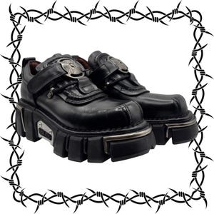 Op de afbeelding: Zwarte leren plateauschoenen met een dikke, grove rubberen zool en metalen accenten. De schoenen zijn voorzien van een klittenbandsluiting over de wreef, een rond metalen embleem op de tong en een zilverkleurige plaat met tekst aan de zijkant.