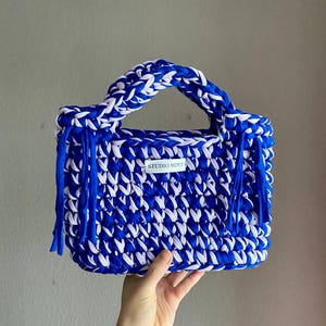 Könnte beinhalten: Eine rechteckige Häkeltasche aus dickem, leuchtend blauem und weißem Stoffgarn. Die Tasche verfügt über zwei stabile Tragegriffe, dekorative Fransen an beiden Seiten und ein kleines weißes rechteckiges Etikett mit der Aufschrift STUDIO MINT.