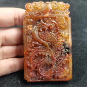 May include: A rectangular, amber-colored pendant with a carved phoenix design. The pendant features intricate details of the bird and floral elements. The phoenix is depicted with outstretched wings and a long tail. The pendant is held in a hand.