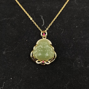 以下が含まれることがあります： 金色のネックレス。翡翠の仏陀ペンダントが付いています。ペンダントは薄緑色で、笑顔の顔があり、金色の縁取りと小さな赤と透明のアクセントで縁取られています。チェーンも金色です。