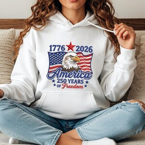 May include: White hoodie featuring a patriotic design. The graphic includes the text "1776 2026 America 250 Years of Freedom" with an American flag and an eagle. The design is in red, white, and blue.