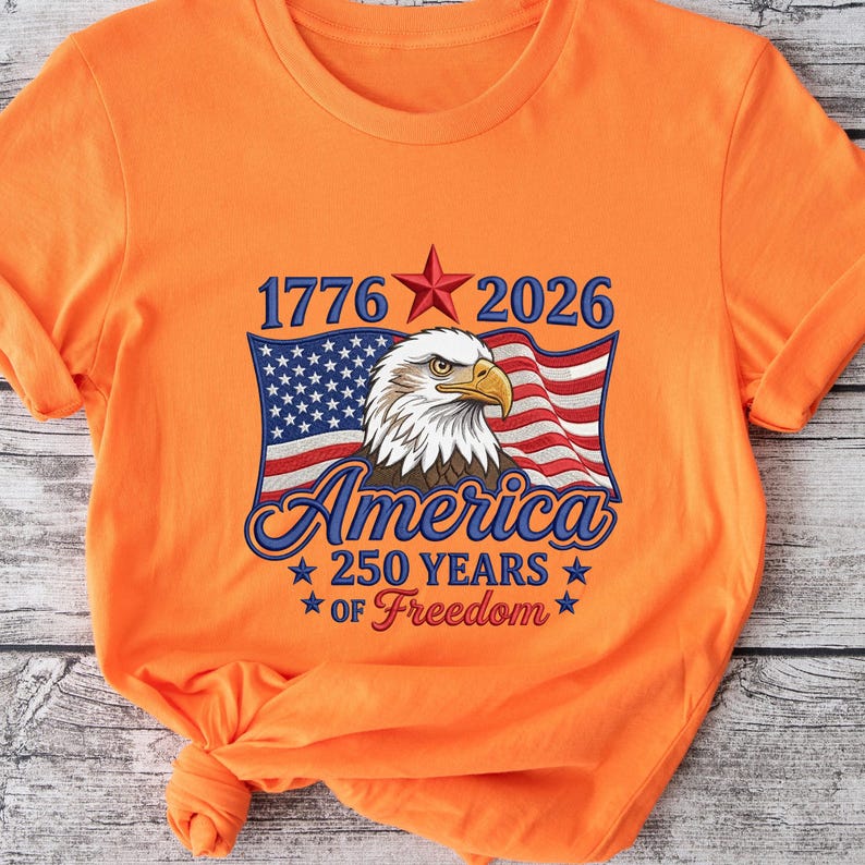 May include: Orange t-shirt featuring a patriotic design. The graphic includes an American flag, a bald eagle, and the text "1776 2026 America 250 Years of Freedom" in blue and red. The shirt is made of a soft material.