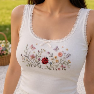 May include: A white ribbed tank top featuring a scoop neckline with delicate lace trim and a small center bow. The chest area is decorated with an embroidered design of a red ladybug surrounded by colorful flowers, green vines, and butterflies.