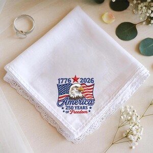 May include: White handkerchief with a delicate lace trim. The handkerchief features a patriotic design with an American flag, an eagle, and the text "America 250 Years of Freedom" with the years 1776 and 2026.