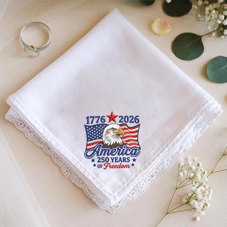 May include: White handkerchief with a delicate lace trim. The handkerchief features a patriotic design with an American flag, an eagle, and the text "America 250 Years of Freedom" with the years 1776 and 2026.