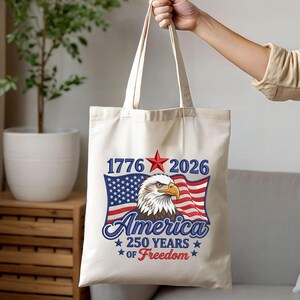 May include: Cream-colored tote bag featuring a patriotic design. The design includes an American flag, an eagle, and the text "1776 2026 America 250 Years of Freedom" in blue and red. The bag is held by a person.