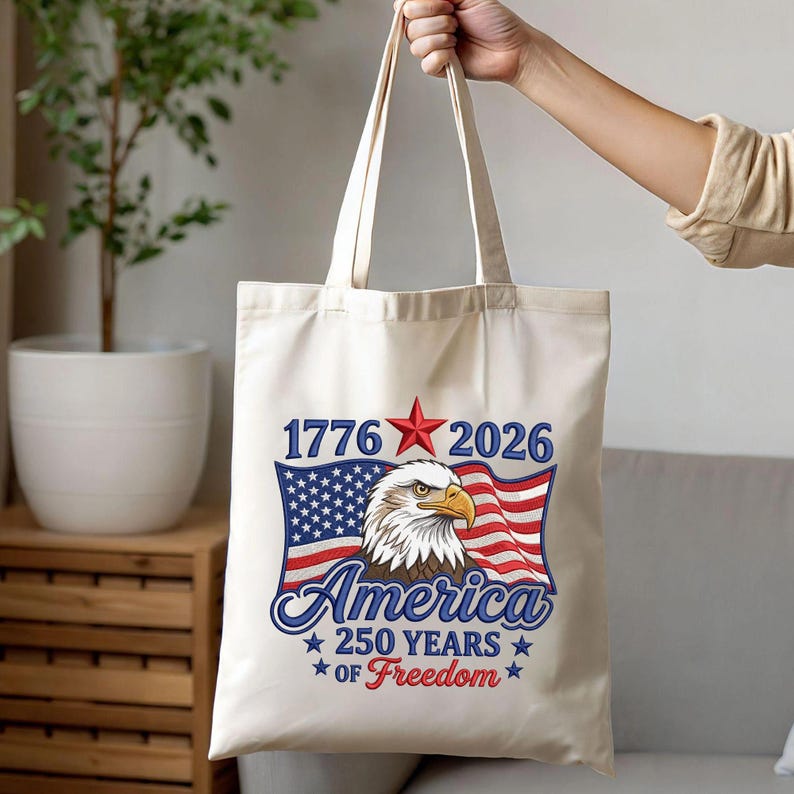May include: Cream-colored tote bag featuring a patriotic design. The design includes an American flag, an eagle, and the text "1776 2026 America 250 Years of Freedom" in blue and red. The bag is held by a person.