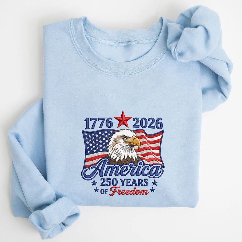 May include: Light blue sweatshirt featuring an American flag design with an eagle and the text "1776 2026 America 250 Years of Freedom". The design includes a red star and patriotic colors.