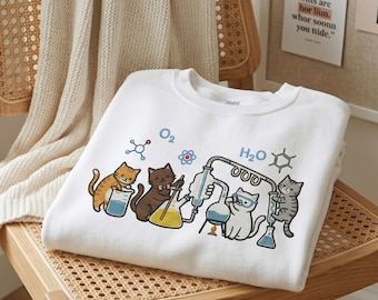 May include: White sweatshirt featuring a science-themed graphic with cartoon cats. The design includes beakers, flasks, and the chemical symbols O2 and H2O. The cats are depicted in various poses, interacting with the lab equipment.