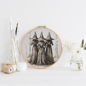 Pode incluir: Um bordado redondo apresenta uma ilustração em aquarela de três figuras com vestidos longos e chapéus pontudos, em um cenário de floresta. A obra de arte é exibida em uma superfície branca, com materiais de arte e um pequeno vaso de flores por perto.