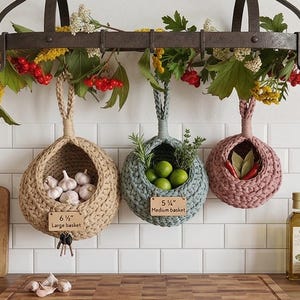 May include: Three hanging crochet baskets in beige, blue, and pink, filled with garlic, limes, and spices. The beige basket is labeled "6 1/2" Large basket". The baskets hang from a metal rack adorned with greenery and berries. Olive oil bottles are in the background.