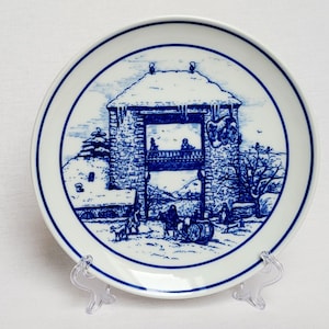Puede incluir: Un plato de cerámica blanco con una ilustración azul y blanca de una escena invernal. El plato presenta una representación detallada de un edificio con personas, árboles y un paisaje nevado. El plato se exhibe sobre un soporte de plástico transparente.