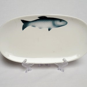 Może przedstawiać: Owalny, kremowy ceramiczny półmisek do serwowania z niebieską ilustracją ryby. Ryba jest wyśrodkowana na półmisku i ma szczegółowy wzór. Półmisek jest podparty przezroczystym plastikowym stojakiem.