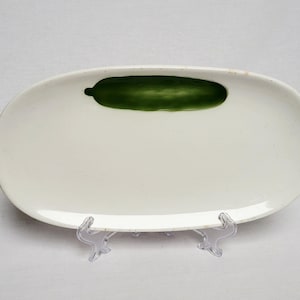 Może przedstawiać: Owalny, kremowy ceramiczny talerz do serwowania z ręcznie malowanym zielonym wzorem ogórka. Talerz jest podparty przezroczystym plastikowym stojakiem, prezentującym prosty, elegancki design. Idealny do serwowania przystawek lub małych potraw.