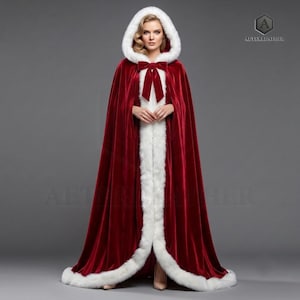 May include: A floor-length deep red velvet cloak featuring a white faux fur trim along the hood, front opening, and hem. The garment includes a matching red velvet bow tie closure at the neck. The logo AETERLEATHER is visible in the top right corner.