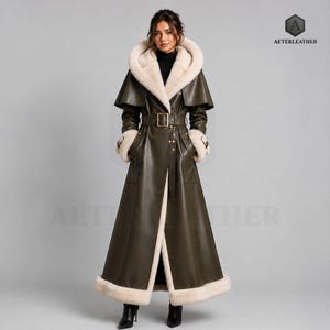 May include: An olive green floor-length leather coat featuring a large hood lined with cream-colored faux fur, a matching faux fur trim along the hem and cuffs, a belted waist with a gold buckle, and a cape-style overlay across the shoulders.