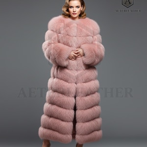 Puede incluir: Un abrigo largo de piel rosa claro con un diseño en capas. El abrigo tiene un cuello ancho y se usa con tacones rosas a juego. El logotipo "AETERLEATHER" es visible en la esquina superior derecha.