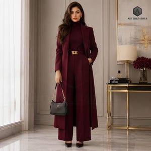 May include: A deep burgundy ensemble consisting of a long, structured coat, a matching turtleneck sweater, and high-waisted wide-leg trousers. The outfit features a gold-toned belt buckle and is paired with a small, black structured handbag.