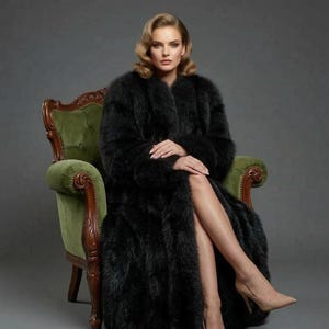 Puede incluir: Una mujer con un abrigo largo de piel sintética negra, sentada en un sillón de terciopelo verde. El abrigo está abierto, revelando las piernas y tacones altos color nude. El fondo es gris liso.