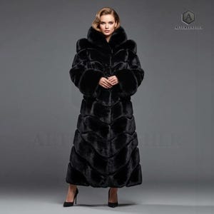 May include: Full-length black faux fur coat with a hooded collar and button closure. The coat has a textured, layered design and is worn with black high heels. The logo "ALTERLEATHER" is visible in the upper right corner.