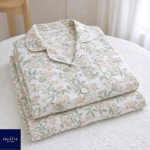 Puede incluir: Un conjunto de pijama floral doblado. El pijama es blanco con un estampado de flores y hojas verdes y beige. La parte superior tiene cuello, botones y un bolsillo en el pecho. El conjunto est&aacute; cuidadosamente apilado sobre una superficie blanca.