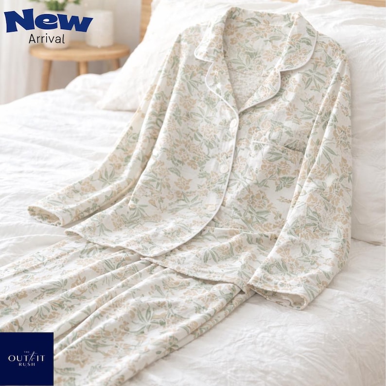 Puede incluir: Un conjunto de pijama de dos piezas con estampado floral en blanco, verde y beige. La parte superior de manga larga tiene botones y un bolsillo en el pecho. Los pantalones son largos y tienen un dise&ntilde;o floral a juego. El texto "New Arrival" es visible.