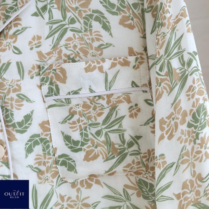 Puede incluir: Primer plano de una camisa de pijama blanca con un estampado floral en tonos verdes y beige. La camisa tiene un bolsillo en el pecho con ribete blanco y una botonadura. El logotipo de Out It Rush es visible en la esquina inferior izquierda.