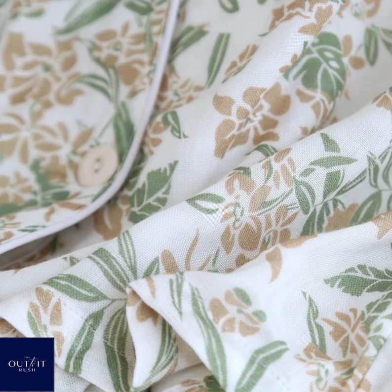Puede incluir: Primer plano de un conjunto de pijama blanco con un estampado floral en tonos verdes y beige. La tela es suave y fluida, con una camisa abotonada y ribete blanco. El logotipo de Outfit Rush es visible.