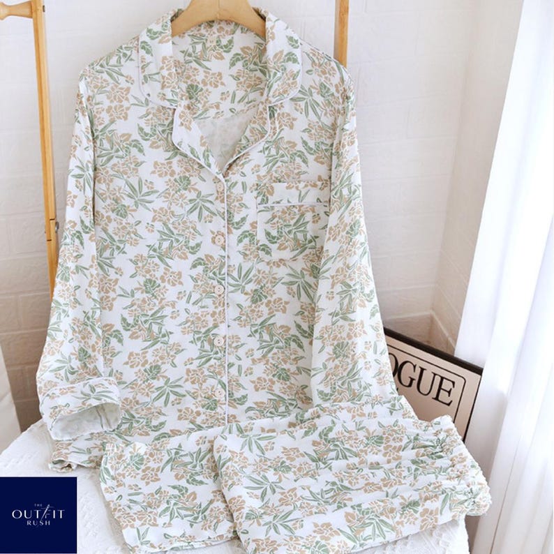 Puede incluir: Un conjunto de pijama blanco con estampado floral en tonos verdes y beige. El conjunto incluye una camisa abotonada con bolsillo en el pecho y pantal&oacute;n a juego. El pijama se exhibe en una percha de madera.