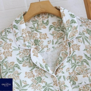 Puede incluir: Camiseta de pijama blanca con estampado floral en tonos verdes y beige. La camiseta tiene cuello, cierre de botones y un peque&ntilde;o bolsillo. El ribete es blanco. La marca "OUT IT RUSH" es visible en la esquina inferior izquierda.