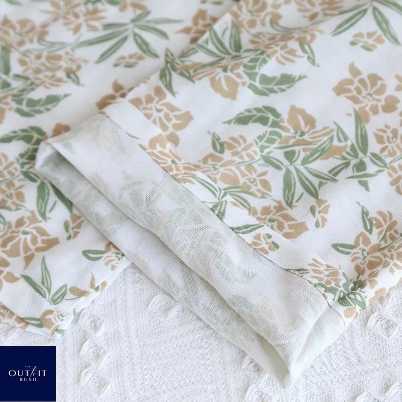 Puede incluir: Primer plano de tela blanca doblada con un estampado floral en tonos verdes y beige. La tela est&aacute; en capas, revelando una impresi&oacute;n detallada. El logotipo "OUT/IT RUSH" es visible en la esquina inferior izquierda.