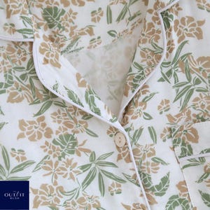 Puede incluir: Primer plano de una camisa de pijama blanca con un estampado floral en tonos verdes y beige. La camisa tiene una botonadura, un cuello con ribete blanco y un peque&ntilde;o bolsillo. La marca "OUT IT RUSH" es visible en la esquina inferior izquierda.