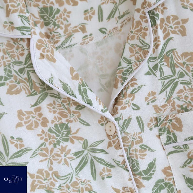 Puede incluir: Primer plano de una camisa de pijama blanca con un estampado floral en tonos verdes y beige. La camisa tiene una botonadura, un cuello con ribete blanco y un peque&ntilde;o bolsillo. La marca "OUT IT RUSH" es visible en la esquina inferior izquierda.