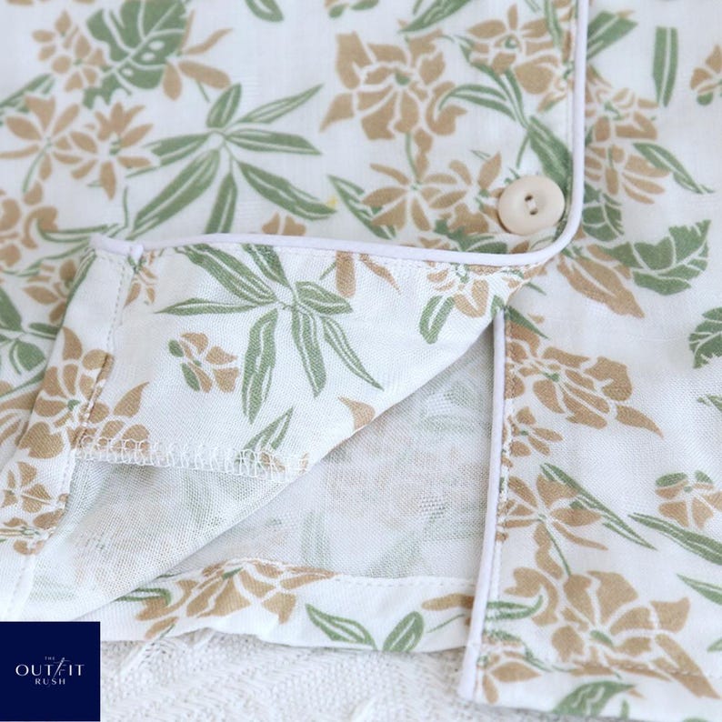 Puede incluir: Primer plano de una camisa de pijama blanca con un estampado floral en tonos verdes y beige. La camisa tiene ribetes blancos y un cierre de bot&oacute;n &uacute;nico. El texto "OUT/IT RUSH" es visible en la esquina inferior izquierda.