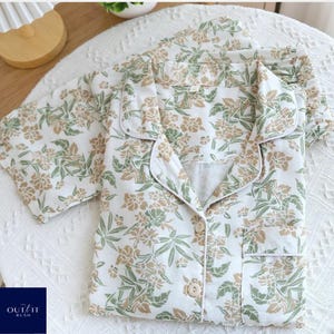 Puede incluir: Conjunto de pijama blanco con estampado floral en tonos verdes y beige. El conjunto incluye una camisa abotonada con bolsillo en el pecho y pantal&oacute;n a juego. Los pijamas son de tejido suave y est&aacute;n sobre una superficie blanca.