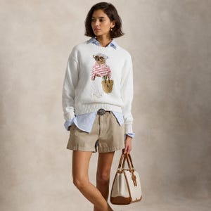 May include: A white long-sleeved sweater featuring a central embroidered bear graphic wearing a striped shirt and hat, layered over a light blue collared shirt, paired with khaki shorts and a white and brown structured handbag with dual handles.