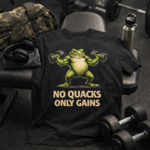 Könnte beinhalten: Schwarzes T-Shirt mit einem muskulösen grünen Frosch, der Hanteln hebt. Darunter steht der Text "NO QUACKS ONLY GAINS". Das Shirt wird vor dem Hintergrund eines Fitnessstudios mit Gewichten und einer Wasserflasche gezeigt.