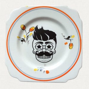 May include: Square white plate featuring a black skull design with star-shaped glasses and a mustache. The plate has an orange and grey border and floral accents. A unique decorative piece for the kitchen or dining room.