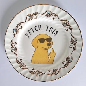 May include: White ceramic plate with a gold rim and decorative brown and teal accents. The plate features a cartoon golden retriever wearing sunglasses and giving the middle finger, with the text "FETCH THIS".