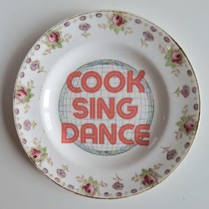 May include: A white ceramic plate with a floral pattern and gold trim. The centre features the words "COOK SING DANCE" in coral-coloured block letters over a blue and white globe design. The plate has a vintage aesthetic.