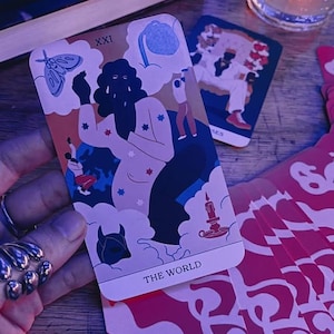 May include: A tarot card from a deck, featuring a central figure with a dark silhouette, surrounded by clouds and stars. The card is labeled "THE WORLD" at the bottom and "XXI" at the top. Other cards and a drink are in the background.